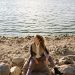A woman sits on a rocky shoreline, gazing at the serene lake view during a sunny day.