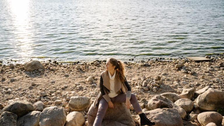 A woman sits on a rocky shoreline, gazing at the serene lake view during a sunny day.